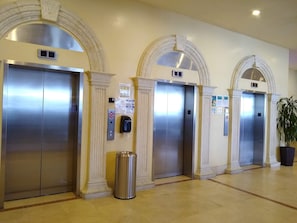 Um saguão de hotel com quatro elevadores, cada um com acabamento metálico, ladeado por colunas e arcos brancos.