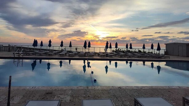 A swimming pool with lounge chairs and umbrellas, reflecting the sunset over the ocean.