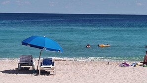A beach with a blue umbrella, lounge chairs, and people swimming and relaxing in the water.