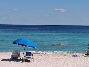 Uma praia com guarda-sol azul, espreguiçadeiras e pessoas nadando e relaxando na água.