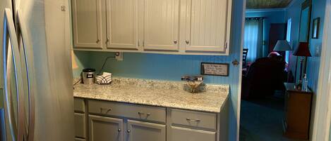 Una cocina con refrigerador de acero inoxidable, encimera de granito y armarios blancos. Hay un cartel en la pared y una lámpara sobre una mesita.