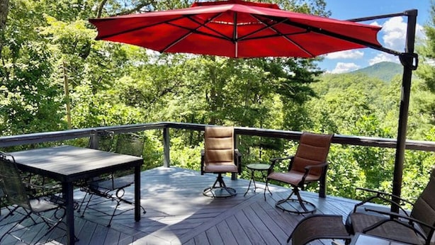 En udendørs terrasse med et bord og stole, en rød parasol og udsigt til et skovområde.
