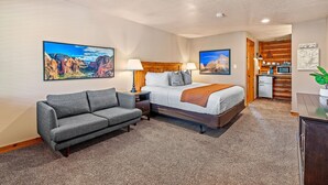 Ein Hotelzimmer mit einem Bett, einem grauen Sofa, einem Nachttisch, einer Lampe, einer Kaffeemaschine und einer kleinen Küchenzeile.