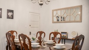 A dining room with a wooden table set for six, surrounded by six wooden chairs. A chandelier hangs from the ceiling, and there are framed pictures on the walls.