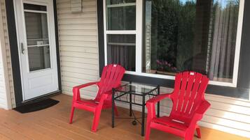 A porch with two red Adirondack chairs, a glass table, and a white door with a window above it.