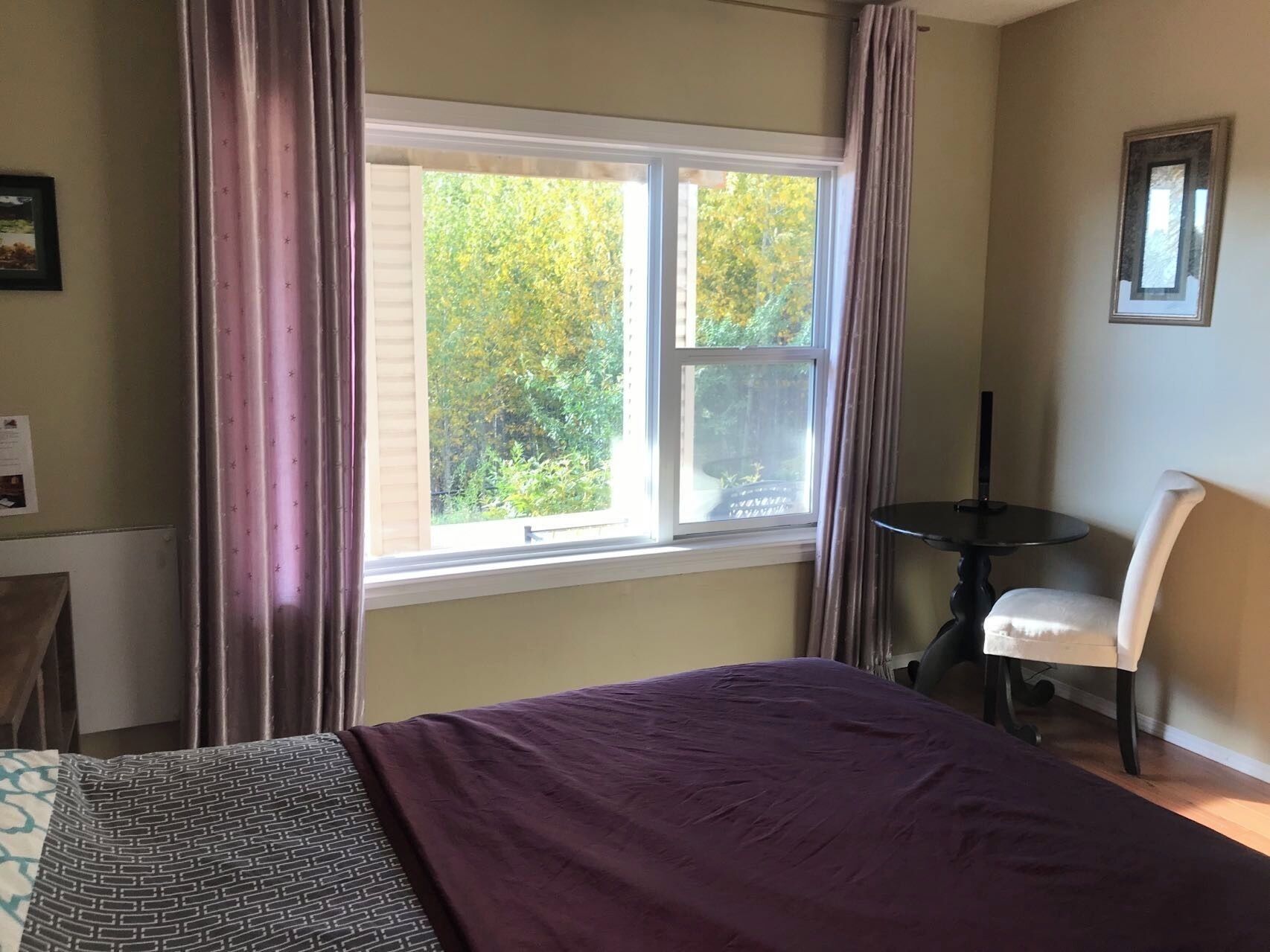 A bedroom with a large window, a bed with a purple bedspread, a small round table, and a chair.