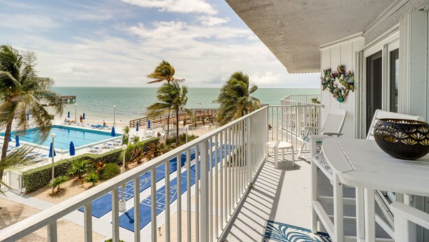 A balcony with a view of a pool and beach, white railings, and a decorative vase.