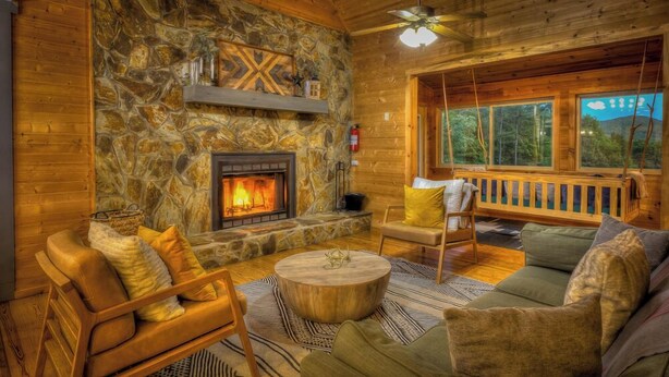 A cozy log cabin interior with a stone fireplace, wooden furniture, and a large window offering a view of the outdoors.