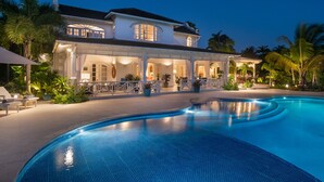 Une grande maison bien éclairée, dotée d’une piscine, entourée de palmiers et agrémentée de mobilier d’extérieur.