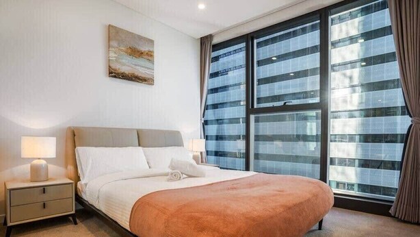 A hotel room with a large window, a bed with a beige headboard, a nightstand with a lamp, and a painting on the wall. - CASSA ONE - Luxury River-View 1 Bed Apt in Brisbane City! (Brisbane)