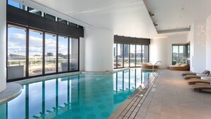 A modern indoor swimming pool with clear glass walls and a view of the cityscape.