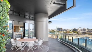 Un espace de restauration extérieur moderne, avec une table ronde et des chaises blanches, offrant une vue sur le bord de l’eau, avec un pont et une grande roue en arrière-plan.