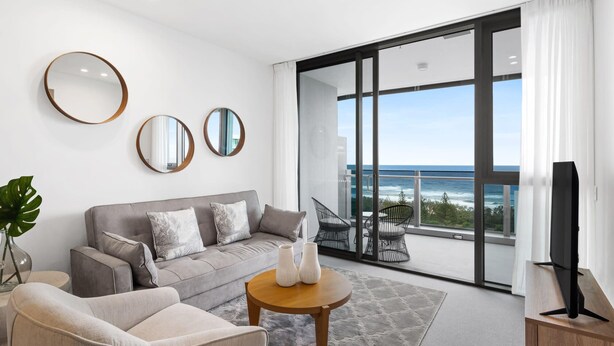 A modern living room with a grey sofa, two armchairs, a coffee table, and a TV on a wooden stand. There are three round mirrors on the wall, and a large window with a view of the ocean.
