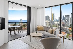 A modern living room with a sofa, coffee table, and chairs, a balcony with a view of the ocean and city skyline.