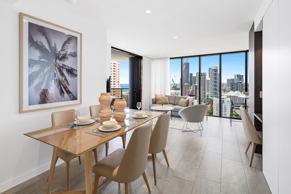 A modern dining area with a city view, a living room with a sofa and a TV, and a balcony with a cityscape.