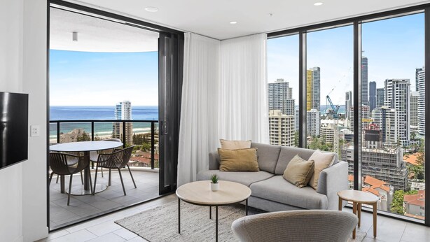 Una sala de estar moderna con sofá, mesa de centro y sillones, un balcón con vista al océano y al horizonte de la ciudad.