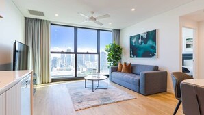 A modern living room with a large window, a ceiling fan, a grey sofa, a coffee table, and a TV. - CASSA ONE - Heart of CBD, City Escape w/ Riverview (Brisbane City)
