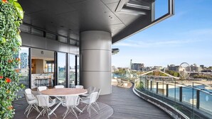 Un área de comedor al aire libre y de diseño moderno, con una mesa redonda y sillas blancas, con vista a un muelle con un puente y una noria al fondo.