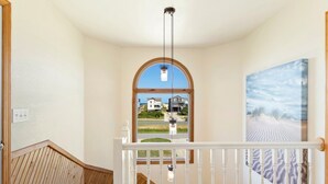 A hallway with a wooden staircase, a large arched window, and a painting of a desert landscape. - 7074 Seashells and Sunsets * 3 Min Walk to Beach (Nags Head)