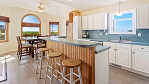 A modern kitchen with a central island, bar stools, and a dining area with a view. - 7074 Seashells and Sunsets * 3 Min Walk to Beach (Nags Head)