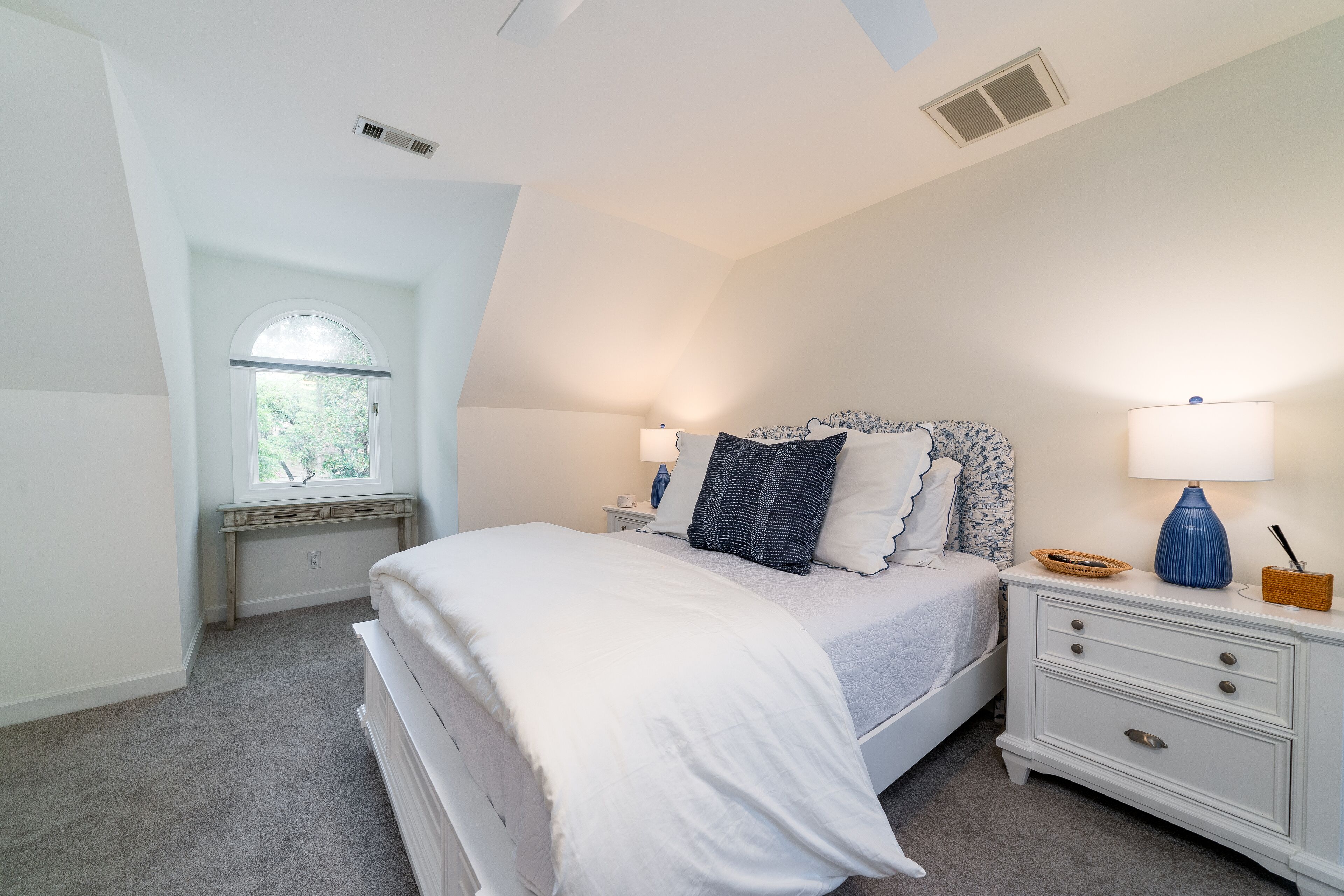 Un dormitorio con cama, mesitas de noche, lámpara, ventana y rejilla en el techo.