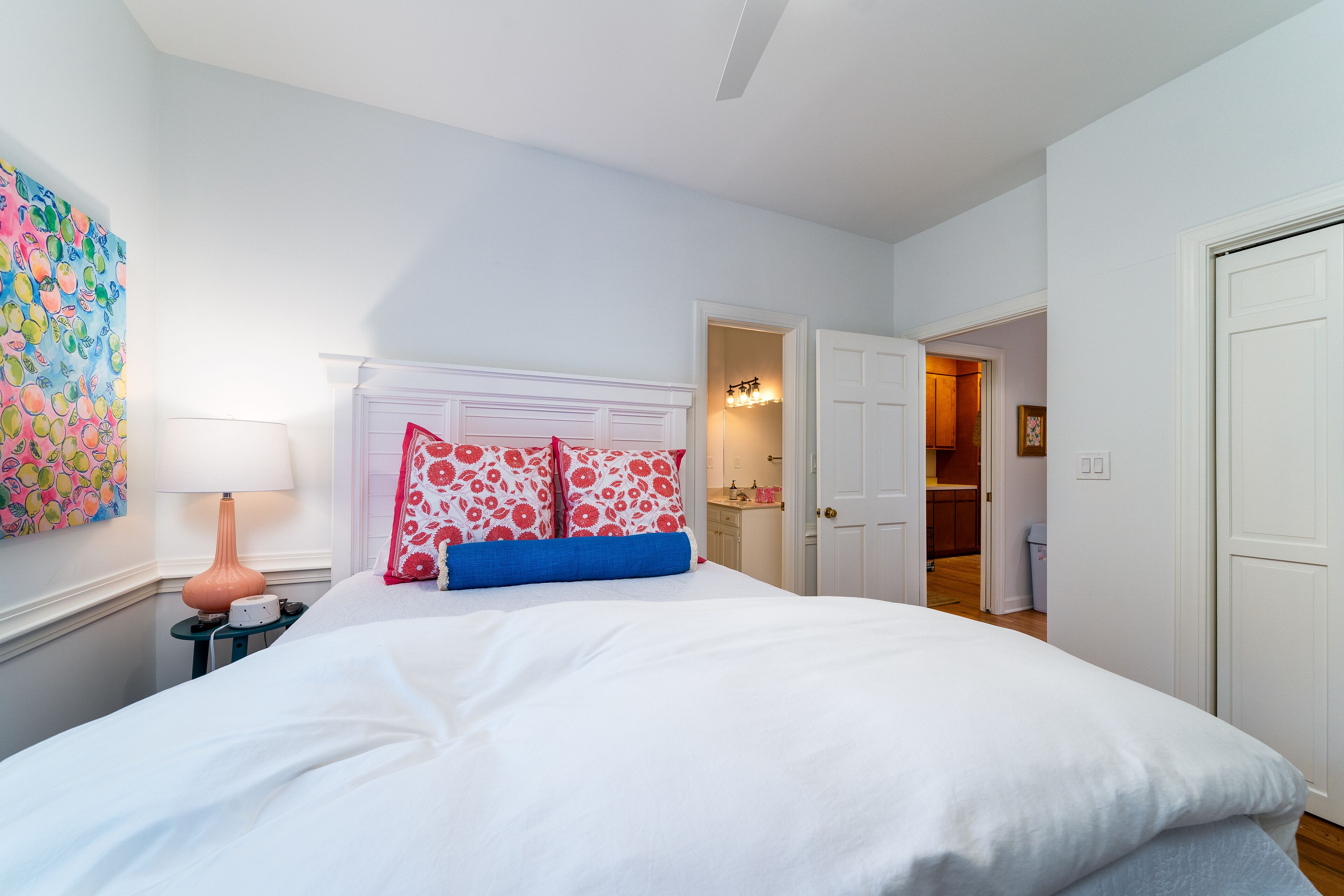 Een slaapkamer met een wit bed, een nachtkastje met een lamp, en een kleurrijke fruitwanddecoratie.