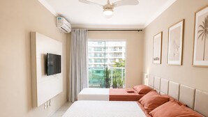 A hotel room with a bed, a ceiling fan, a TV, a window with a view of buildings, and a potted plant. - Confortável Apto na Praia do Forte (Cabo Frio)