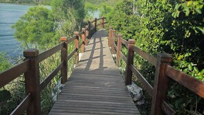 A wooden boardwalk leading to a body of water, surrounded by greenery and hills. - Confortável Apto na Praia do Forte (Cabo Frio)