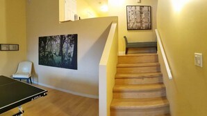 A room with a ping pong table, a staircase with a wooden finish, a framed painting of a forest, and a white chair. - Grant's Arctic Escape (Iqaluit)