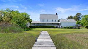 A white house with a porch, a wooden dock leading to it, and a grassy field. - Oyster Catcher | Deep Creek Waterfront w/ Deep Water Dock, Screened Porch, Guest Cottage & Pets (Chincoteague Island)