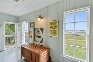 A room with a wooden bench, a wall-mounted light fixture, a framed poster, and a large window with a view of the outdoors. - Oyster Catcher | Deep Creek Waterfront w/ Deep Water Dock, Screened Porch, Guest Cottage & Pets (Chincoteague Island)