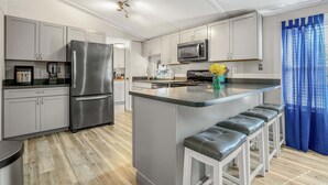 A modern kitchen with a stainless steel refrigerator, a microwave, and a coffee maker. There is a bar area with stools and a vase of yellow flowers. - Counting Corks | Waterfront Pet Friendly Cottage w/ Dock for Crabbing, Firepit & Near Cape Charles (Cape Charles)