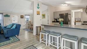 A modern kitchen with a central island, white cabinetry, stainless steel appliances, and a dining area with a blue sofa and bar stools. - Counting Corks | Waterfront Pet Friendly Cottage w/ Dock for Crabbing, Firepit & Near Cape Charles (Cape Charles)