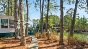 A wooden deck leading to a cabin with a green roof, surrounded by tall trees and a body of water. - Counting Corks | Waterfront Pet Friendly Cottage w/ Dock for Crabbing, Firepit & Near Cape Charles (Cape Charles)