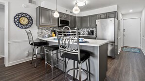 A modern kitchen with a central island, bar stools, stainless steel appliances, and a decorative wall clock. - Chic & charming 2 bedroom oceanfront condo w/amazing views & large pool! (North Myrtle Beach)