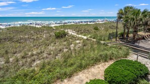 A beach with a sandy path leading to the water, palm trees, and a fenced area with beach umbrellas. - Chic & charming 2 bedroom oceanfront condo w/amazing views & large pool! (North Myrtle Beach)