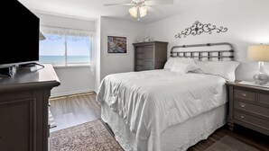 A bedroom with a bed, a nightstand, a dresser, a ceiling fan, a TV, and a window with a view of the ocean. - Chic & charming 2 bedroom oceanfront condo w/amazing views & large pool! (North Myrtle Beach)