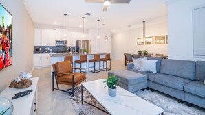 Uma sala de estar moderna com um sofá cinzento, uma mesa de centro e um espaço de refeições com cadeiras, com uma cozinha ao fundo.