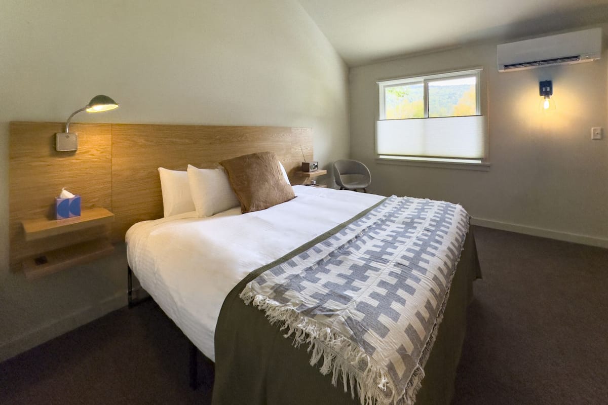 A bedroom with a bed, a nightstand, a lamp, a chair, a window, and an air conditioning unit.