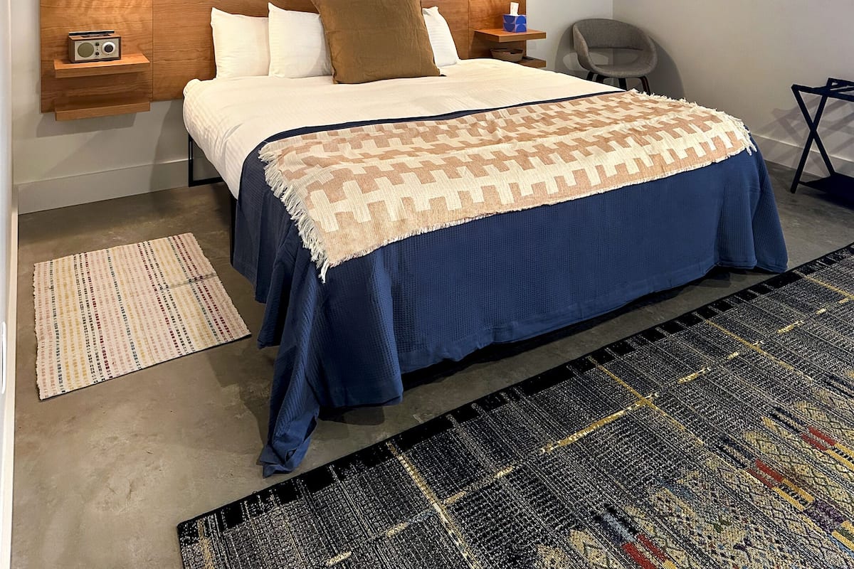 A neatly made bed with a blue bedspread and a patterned throw, flanked by two bedside lamps and a radio on a wooden shelf.