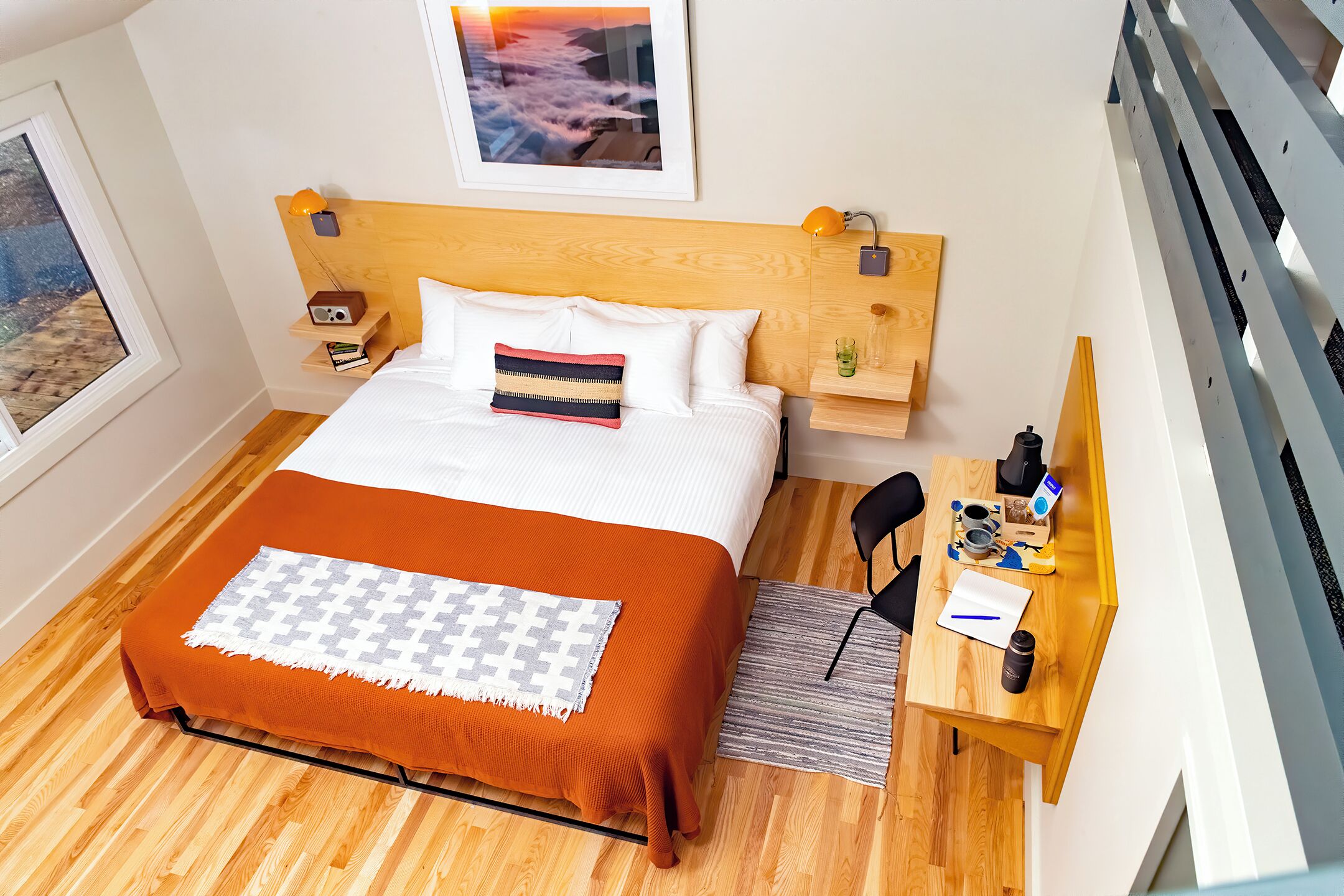 Ein ordentlich eingerichtetes Schlafzimmer mit Holz-Kopfteil, einem Bett mit weißer und orangefarbener Bettwäsche, einem Nachttisch mit Lampe sowie einem Schreibtisch mit Stuhl.