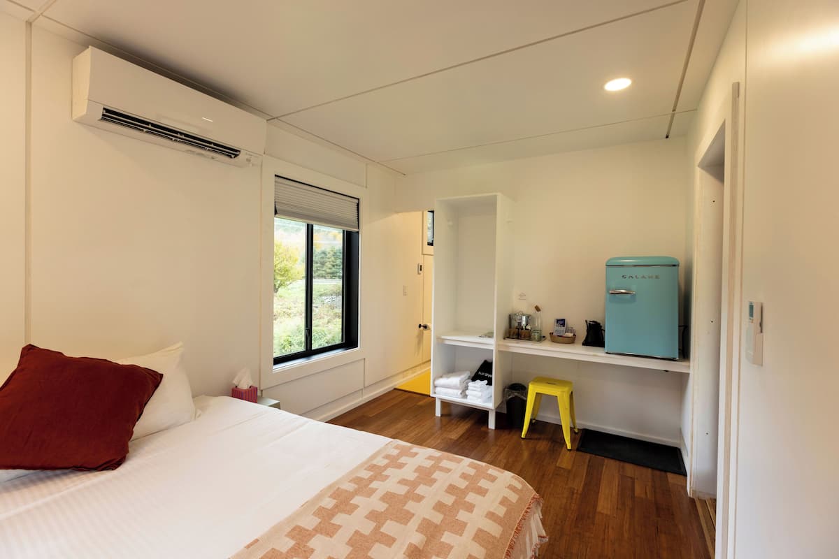 A hotel room with a bed, a window, a wardrobe, a small stool, and a teal-colored refrigerator.