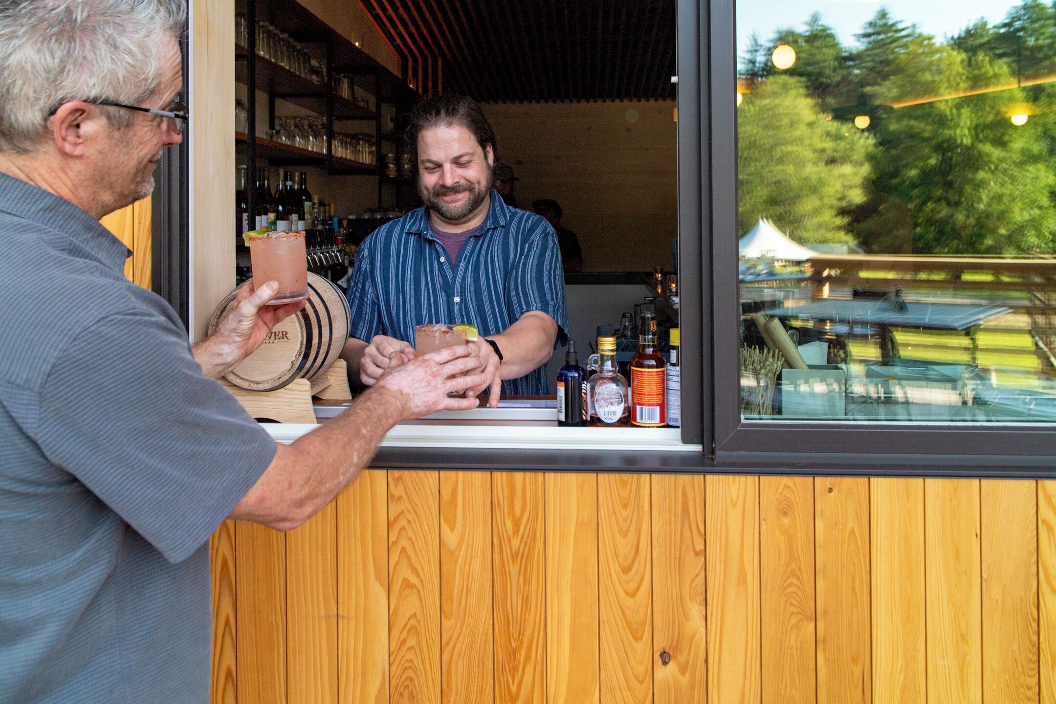Un comptoir de bar avec un revêtement en bois, un barman préparant une boisson, et une sélection de bouteilles et de verres.