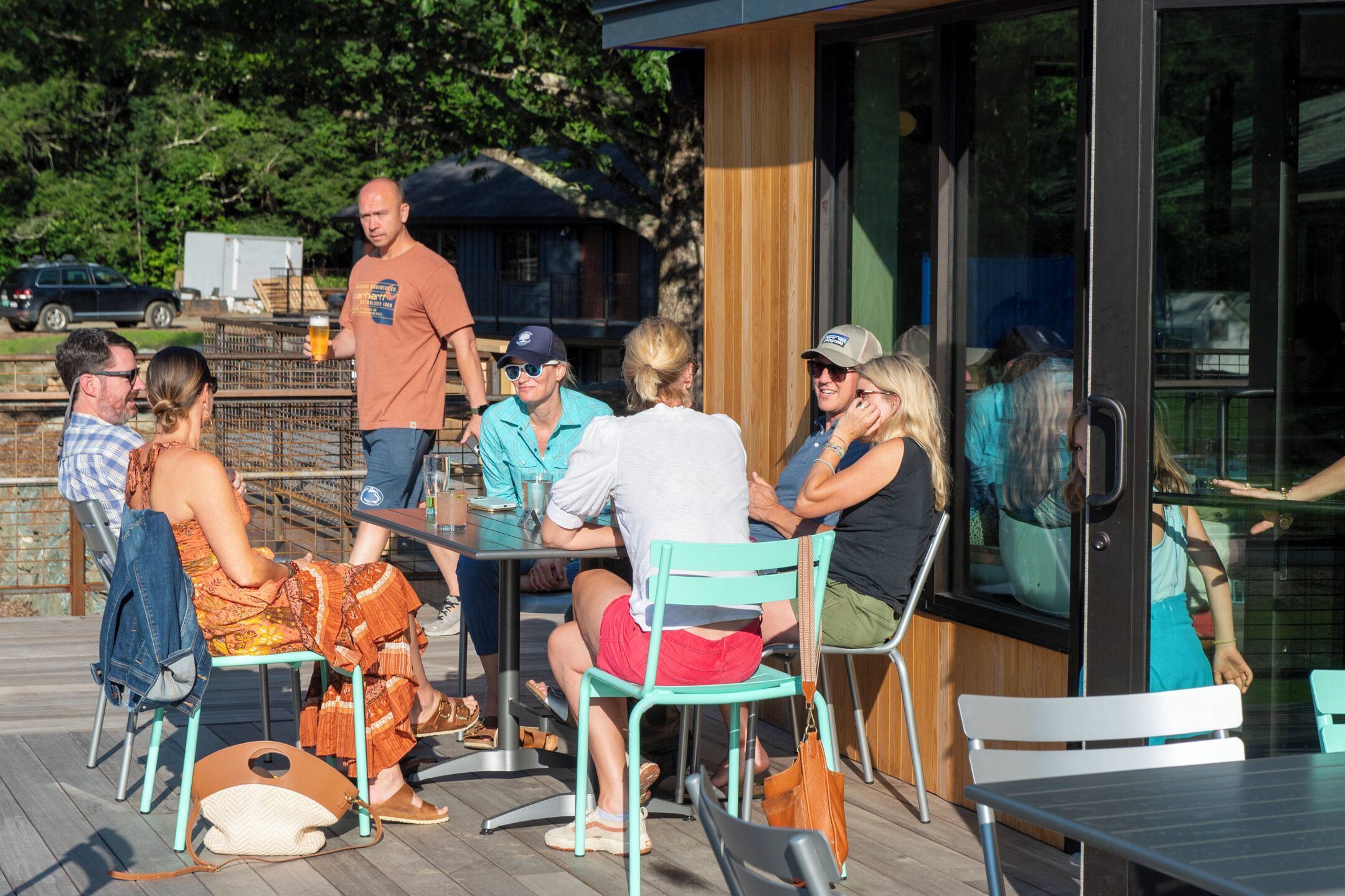A group of people seated at an outdoor table on a wooden deck, with a glass enclosure and metal chairs.