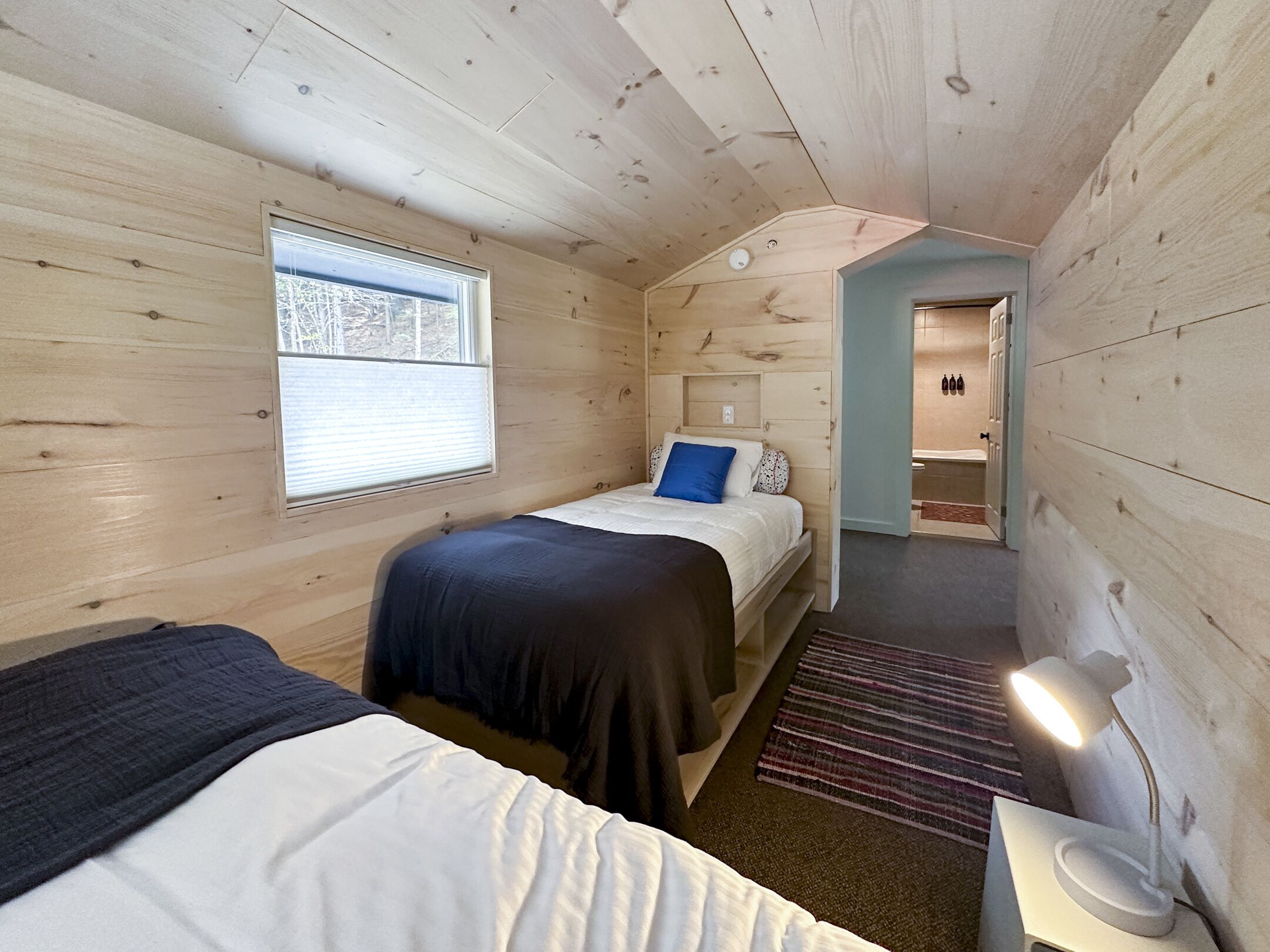 Et hyggeligt værelse i sommerhusstil med to senge, et vindue, en dør til et badeværelse og et lille bord med en lampe.