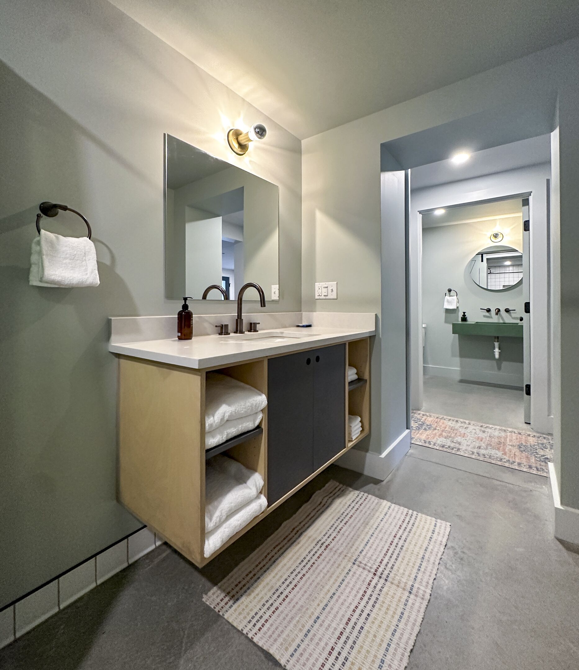 Un bany modern amb un mirall gran, un lavabo i un portaToalletes. Hi ha un armari de fusta amb prestatges per a tovalloles.