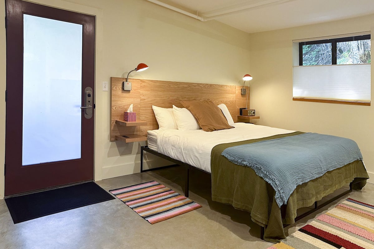 A bedroom with a bed, bedside lamps, a wooden headboard, a window with blinds, and a door with a peephole.