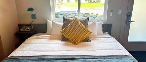 A hotel room with a bed, a window with a view of a park, a nightstand with a radio, and a door with a handle.