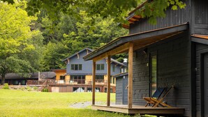 A modern wooden cabin with a covered porch and a deck, surrounded by lush greenery and other cabins. - Madbush Falls (Waitsfield)