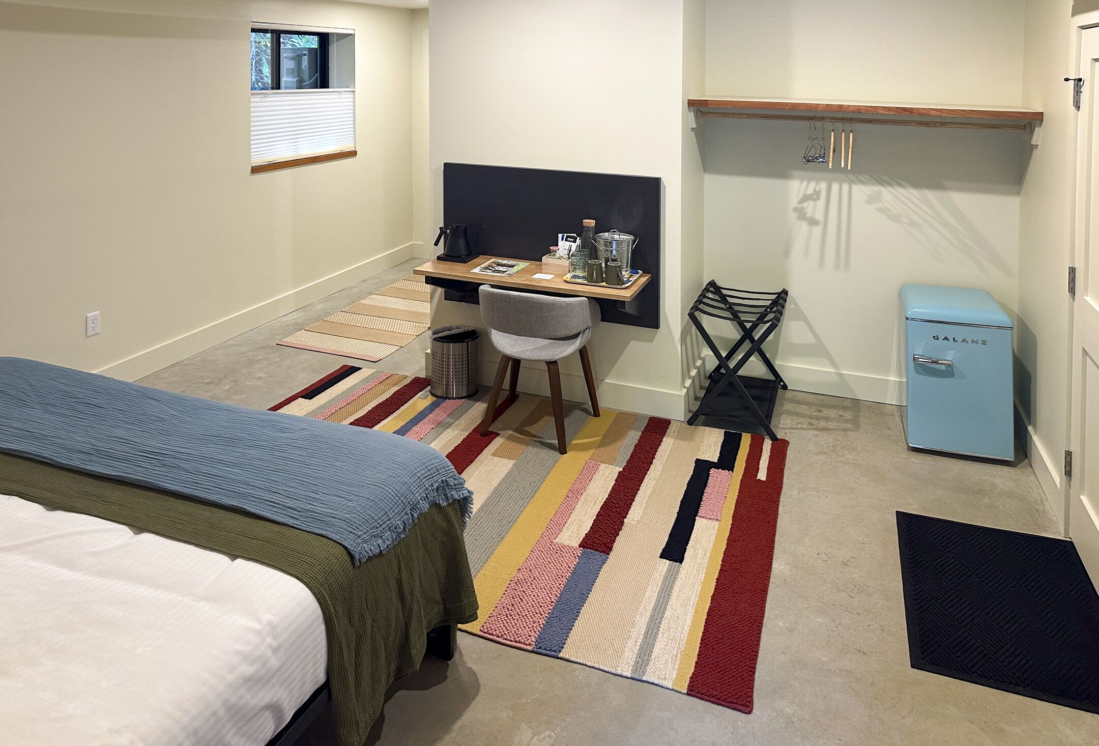 A compact hotel room with a bed, a desk with a chair, a small refrigerator, and a window.